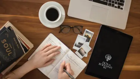 A person's hand journaling on a wooden table with a cup of coffee and a laptop.