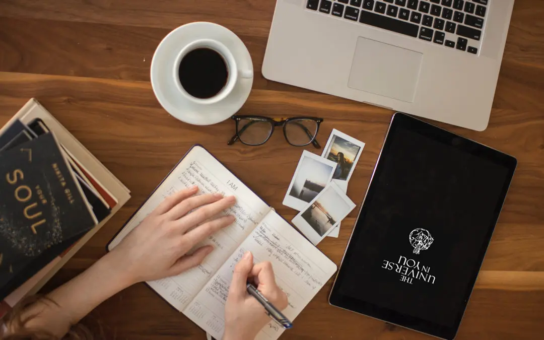 A person's hand journaling on a wooden table with a cup of coffee and a laptop.