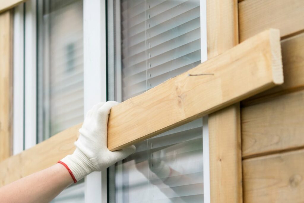 A person wearing gloves nails a plank of wood in front of a window to protect from severe weather. 