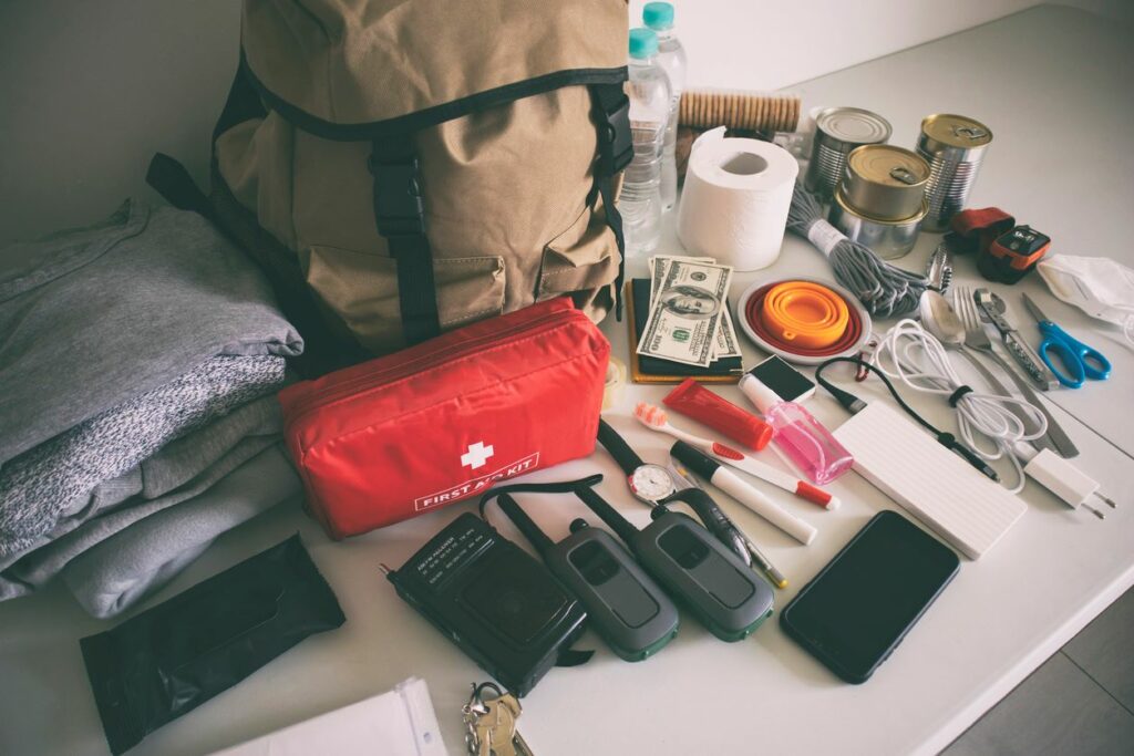 An assortment of items for an evacuation kit, including a first aid kit, walkie-talkies, canned food, charging cables, and more. 